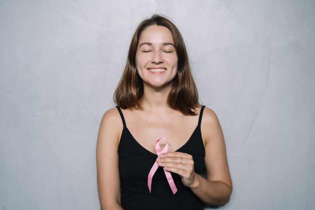 A smiling woman with eyes closed holds a pink breast cancer awareness ribbon, symbolizing hope and health.